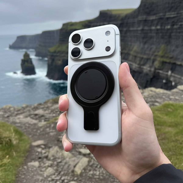 A hand holds a smartphone with a black circular grip stand against a coastal cliff backdrop.