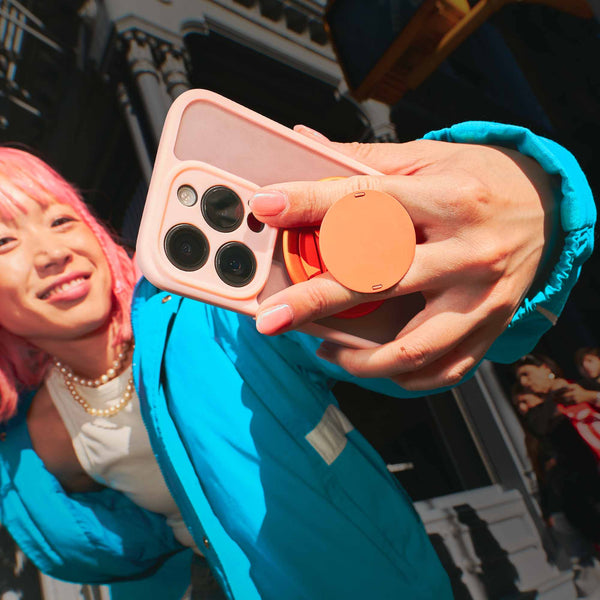 A person with pink hair holds a smartphone with a pink case and a circular grip, wearing a blue jacket.