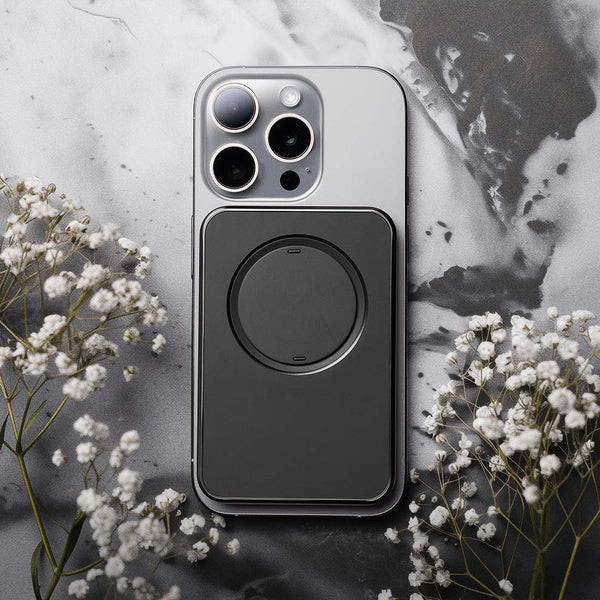 Black snap grip wallet attached to a smartphone on a textured surface with small white flowers nearby.
