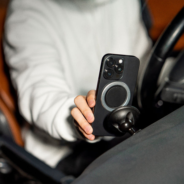 A hand holds a smartphone with a magnetic mount attached, positioned in a car interior.