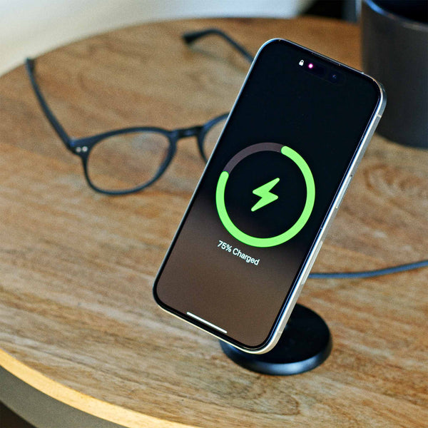 Smartphone displaying a charging status of 75% on a wireless charging stand, with glasses and a mug in the background.