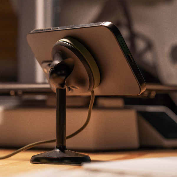 A smartphone mounted on a black stand with a charging cable attached, placed on a wooden desk.