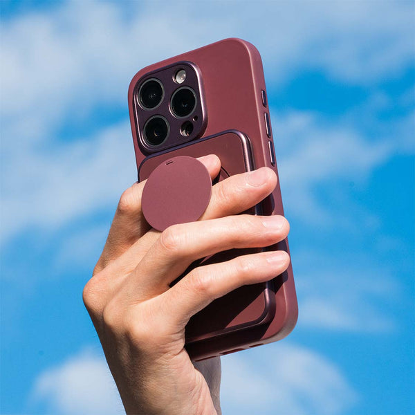 Hand holding a smartphone with a burgundy case and a circular grip accessory against a blue sky.