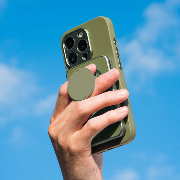 Hand holding a smartphone with a green case and a circular grip against a blue sky.