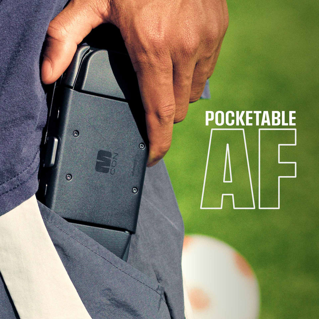 A hand holds a black device partially tucked into a pocket, with grass and a soccer ball visible in the background.
