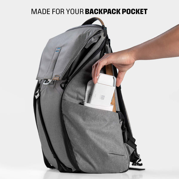 A hand places a white device into the side pocket of a gray backpack.